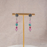 Twee zilverkleurige oorhangers met roze en blauwe natuurstenen, hangend aan een gouden oorbellenstandaard
