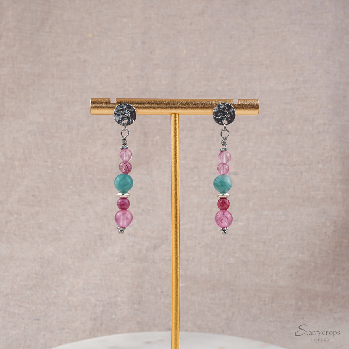 Twee zilverkleurige oorhangers met roze en blauwe natuurstenen, hangend aan een gouden oorbellenstandaard