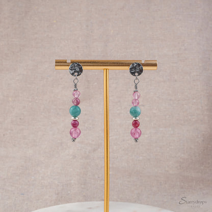 Twee zilverkleurige oorhangers met roze en blauwe natuurstenen, hangend aan een gouden oorbellenstandaard