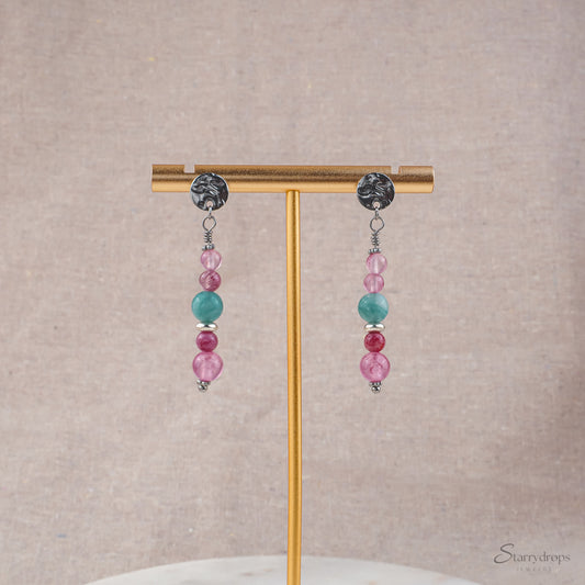 Twee zilverkleurige oorhangers met roze en blauwe natuurstenen, hangend aan een gouden oorbellenstandaard