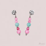Zilverkleurige oorbellen met gehamerd stekertje en kralen in roze, blauw en fuchsia, op een beige achtergrond