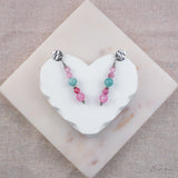 Zilverkleurige oorhangers met roze, blauwe en fuchsia natuurstenen, op een wit hartvormig display met stoffen achtergrond