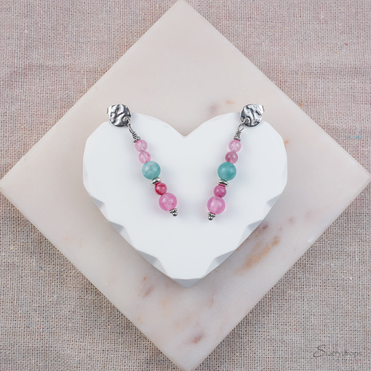 Zilverkleurige oorhangers met roze, blauwe en fuchsia natuurstenen, op een wit hartvormig display met stoffen achtergrond