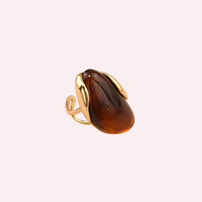 Goudkleurige ring met grote bolle bruine harsknop met amber tinten, liggend op een lichte ondergrond